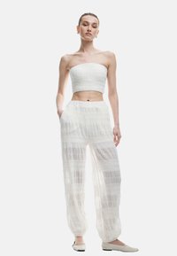 Weißes trägerloses Crop-Top mit strukturiertem Smocking, kombiniert mit lockeren, transparenten weißen Hosen mit horizontalen Streifen und elastischen Bündchen.