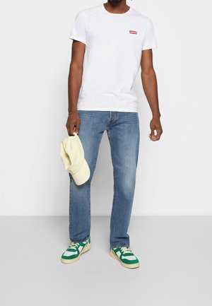 Camiseta blanca de manga corta con logo rojo, jeans de mezclilla azules, zapatillas verdes y blancas, y una gorra de béisbol amarilla sostenida en la mano.