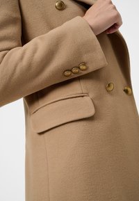 Abrigo beige de doble botonadura con tejido texturizado, cuello con muesca, bolsillo con solapa y botones dorados en las mangas.