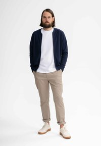 Marineblauer Reißverschluss-Pullover über einem weißen T-Shirt, kombiniert mit hochgekrempelten Khaki-Hosen und weißen Sneakers mit beigen Sohlen. Einfaches, lässiges Design.