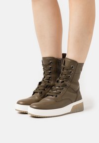 Bottines en cuir vert olive avec lacets sur le devant, fermeture éclair sur le côté, semelle blanche, talon texturé et détail de renforcement à l'orteil.