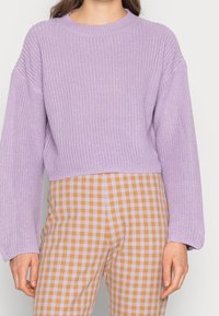 Maglione a coste lilla con scollatura tonda e spalle basse, abbinato a pantaloni a quadretti gialli e lavanda.