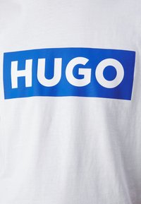 Tricou alb din bumbac cu un grafic rectangular mare de culoare albastră, având cuvântul "HUGO" în litere albe, îngroșate.