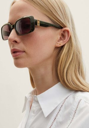 Lunettes de soleil en forme carrée avec des verres foncés et une monture verte, mettant en avant des accents en métal doré. Portées avec une chemise blanche à col.