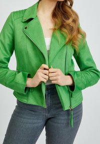 Donna che indossa una giacca zip-up verde brillante sopra una maglietta bianca e jeans grigi scuri, mentre tiene la cerniera della giacca con entrambe le mani.
