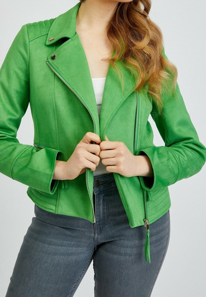 Donna che indossa una giacca zip-up verde brillante sopra una maglietta bianca e jeans grigi scuri, mentre tiene la cerniera della giacca con entrambe le mani.