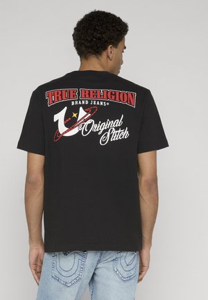 Uomo che indossa una T-shirt nera marchiata True Religion con logo e scritte rosse e bianche sul retro, abbinata a jeans azzurri.