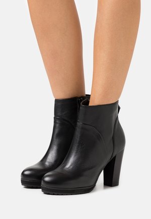 Bottines en cuir noir à talons carrés portées sur des jambes nues contre un fond blanc.