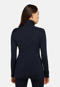 Femme aux longs cheveux noirs portant un pull col roulé ajusté bleu marine, vue de dos sur un fond uni.