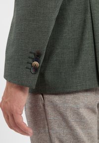 Nahaufnahme eines Arms eines Mannes in einer strukturierten grünen Jacke, die vier Knöpfe am Bund zeigt, kombiniert mit hellbraunen karierten Hosen.