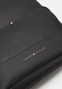 Panel negro texturizado y suave con el logotipo de Tommy Hilfiger y una pequeña etiqueta de tela roja, blanca y azul en un bolso o accesorio.