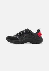 Black athletic shoe featuring mesh and suede materials, a thick sole, curved design, reflective accents, and a red branded heel tab.