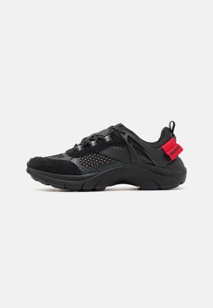 Zwarte sportieve schoen met mesh- en suede-materialen, een dikke zool, een gebogen ontwerp, reflecterende accenten en een rode merklogo-hielstrip.
