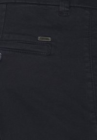 Pantaloni in denim scuro con una texture liscia. Presentano una tasca anteriore, un piccolo dettaglio del logo metallico e accenti di cucitura sottili.