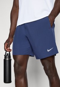 Ναυτικό μπλε σορτσάκι Nike φτιαγμένο από ελαφρύ ύφασμα με πλευρική τσέπη και λευκό λογότυπο swoosh. Μπουκάλι νερού σε μαύρο χρώμα κρατημένο σε ένα χέρι.