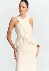 Robe sans manches crème avec un décolleté croisé, un peplum à volants et une jupe ajustée. Texture lisse, design minimal et silhouette élégante.