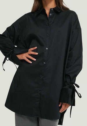 Czarna koszula na długi rękaw z guzikami i zawiązywanymi mankietami, noszona bez wpuszczania w szare spodnie, z ręką opartą na biodrze, na neutralnym tle.