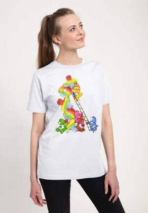 Młoda kobieta w białym T-shircie z kolorowymi postaciami z Care Bears wspinająca się po drabinie na tle dużych liter "TOY" w kolorach tęczy.