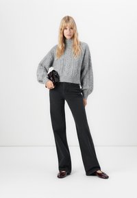 Blonde Frau trägt einen grauen grob gestrickten Pullover, schwarze weite Jeans, bordeauxfarbene Slipper und hält eine kleine dunkle Handtasche vor einem weißen Hintergrund.
