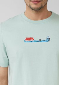 Jasnozielona bawełniana koszulka z haftowanym logo "JAWS" przedstawiającym rekina i fale w niebieskim i białym kolorze, umieszczonym na środku.