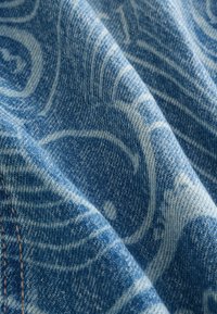 Tissu en denim bleu avec motif paisley blanc et couture visible le long du bord dans un gros plan montrant la texture et les plis.