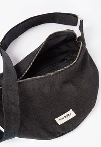 Sac bandoulière en tissu gris foncé avec une fermeture zippée en haut, une poche intérieure et un patch avec une étiquette. Texture lisse, design simple, sangle réglable.
