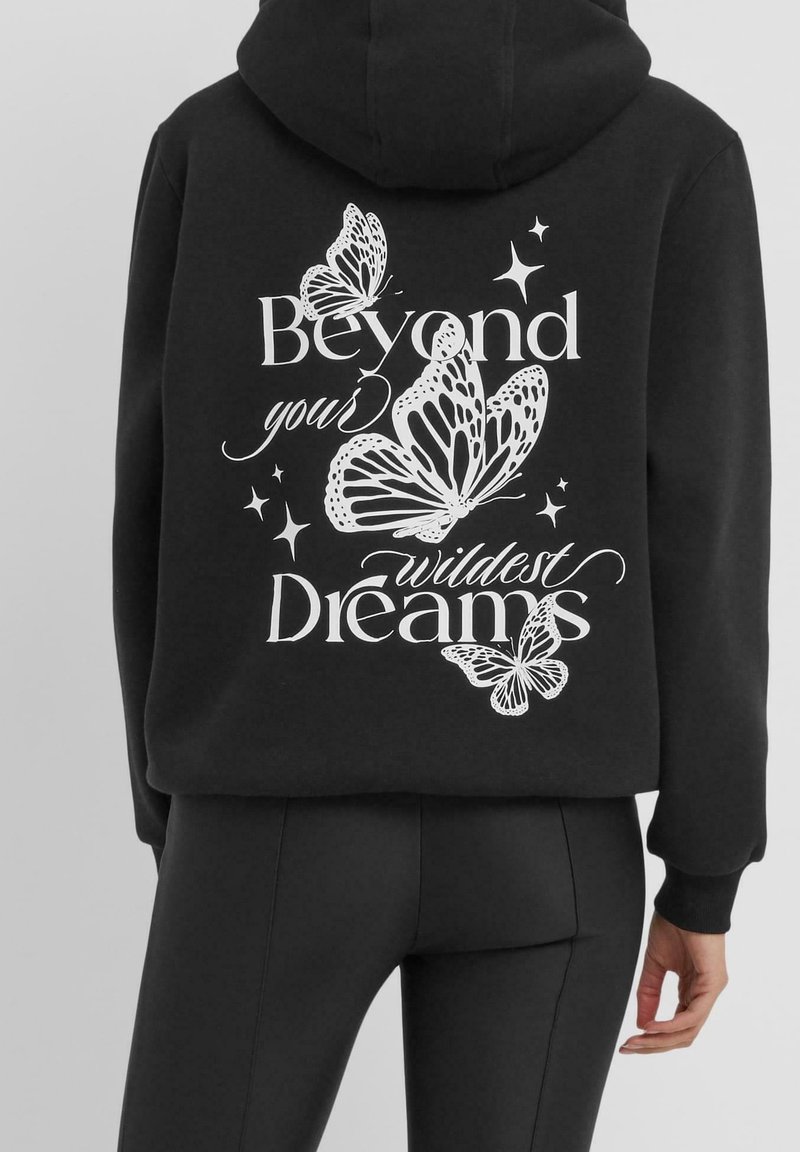 Sweat à capuche noir avec des papillons blancs et des étoiles, avec le texte « Beyond your wildest Dreams » dans le dos, porté avec un pantalon noir.