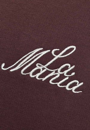 Hnedá tkanina s bielym vyšívaným textom „La Manía“ v plynulom písme. Materiál pôsobí hladko so slabou textúrou.