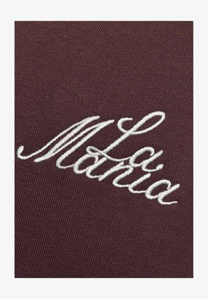 Pruun kangas, millel on valge tikandiga kiri "La Manía" voolavas kirjas. Materjal näib sile, kuid kergelt tekstuuriga.