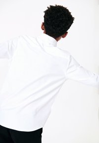 Camisa blanca de manga larga con botones, cuello estructurado y textura suave. Combinada con pantalones negros, vista desde atrás.