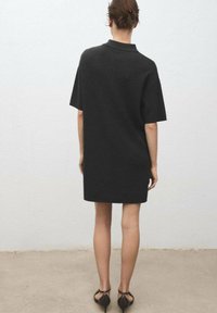 Black oversized shirt dress with a polo collar, short sleeves, and a straight cut, made from a soft material. Thin, strappy black shoes.