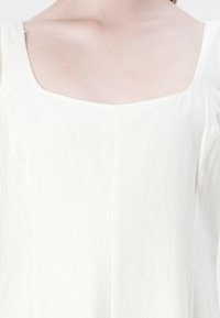 Hvit linkjole med firkantet halslinning og brede stropper. Stoffet har en subtil tekstur og har en vertikal sømdetalj foran.