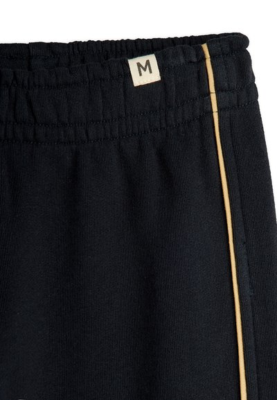 Schwarze Hose mit elastischem Bund, einem kleinen beigen Größenetikett "M" und einem dünnen goldenen vertikalen Streifen auf der rechten Seite.