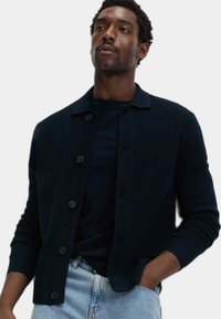 Dunkelblauer Cardigan aus geripptem Strickstoff, mit Knopfverschluss, umgeschlagenem Kragen und langen Ärmeln. Getragen über einem schwarzen Hemd.