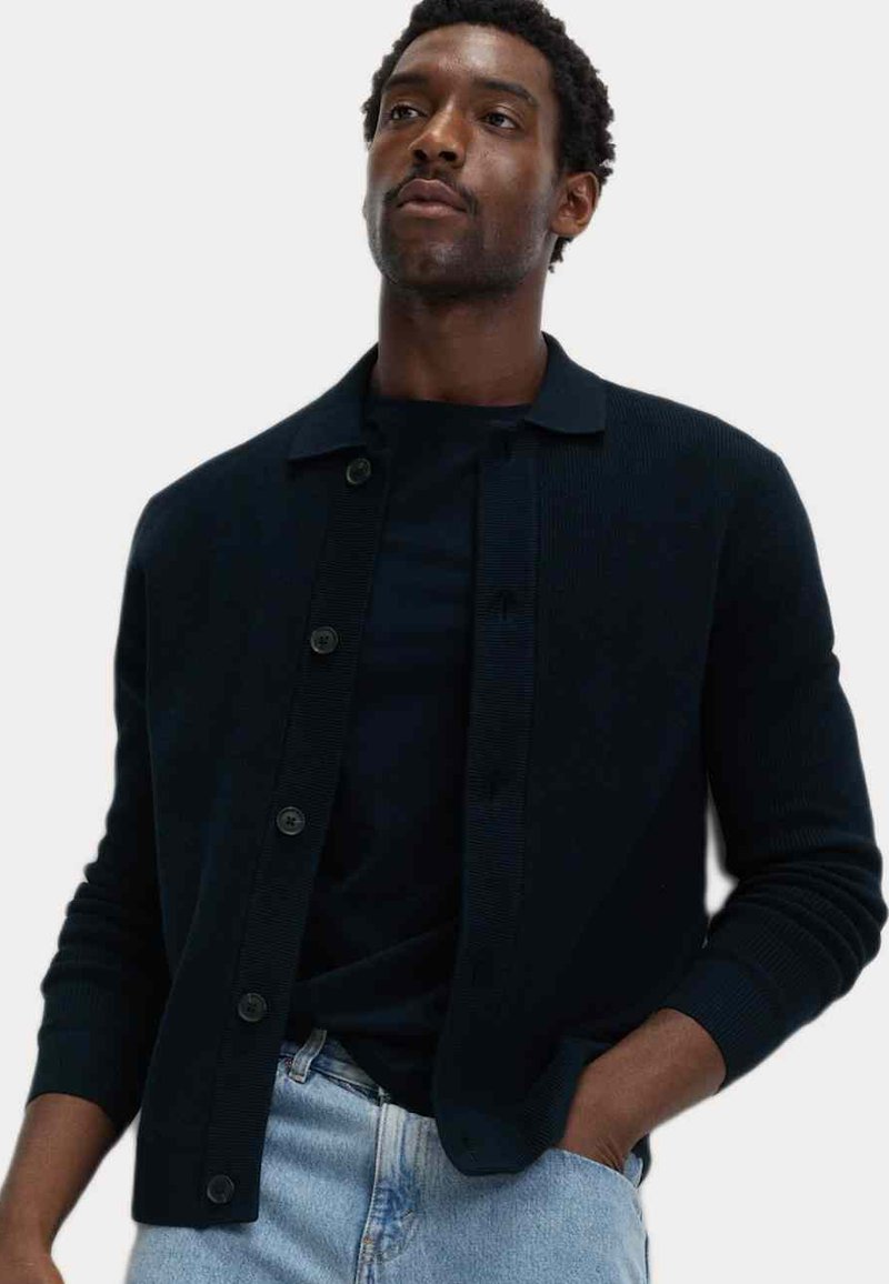 Dunkelblauer Cardigan aus geripptem Strickstoff, mit Knopfverschluss, umgeschlagenem Kragen und langen Ärmeln. Getragen über einem schwarzen Hemd.