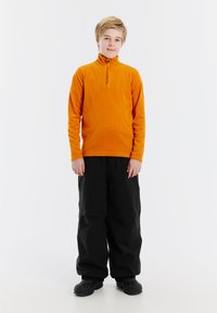 Oranje fleece pullover met hoge kraag en halve rits, gecombineerd met losse zwarte sneeuwbroek en donkere schoenen. Eenvoudig ontwerp, geen patronen.