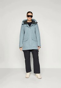 Nais, kes kannab helesinist kapuutsi talvepraktikat, musti pükse, beeže jalanõusid ja suuri musti prille, seisab ühetoonilise tausta taustal.