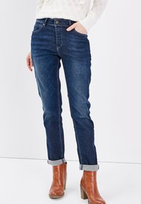 Jean en denim bleu foncé avec une coupe slim, présentant des poches avant et un ourlet roulé. Portés avec des bottines à talons marron.