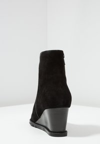 Bottine compensée en daim noir avec une finition mate et lisse, une forme légèrement effilée, et une semelle en caoutchouc noire. Détail décoratif avec une fermeture éclair au dos.