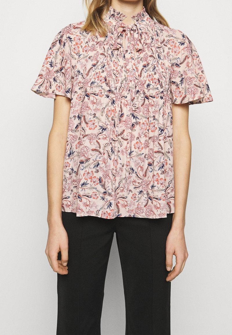 Květinová blůza v světlé růžové barvě, s nařaseným výstřihem, krátkými rukávy a složitými květinovými vzory v tmavě růžové a námořnické modré.