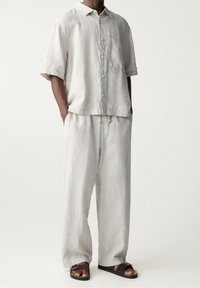 Homme portant une chemise boutonnée à manches courtes en lin léger beige clair et un pantalon large assorti, avec des sandales à enfiler marron foncé.