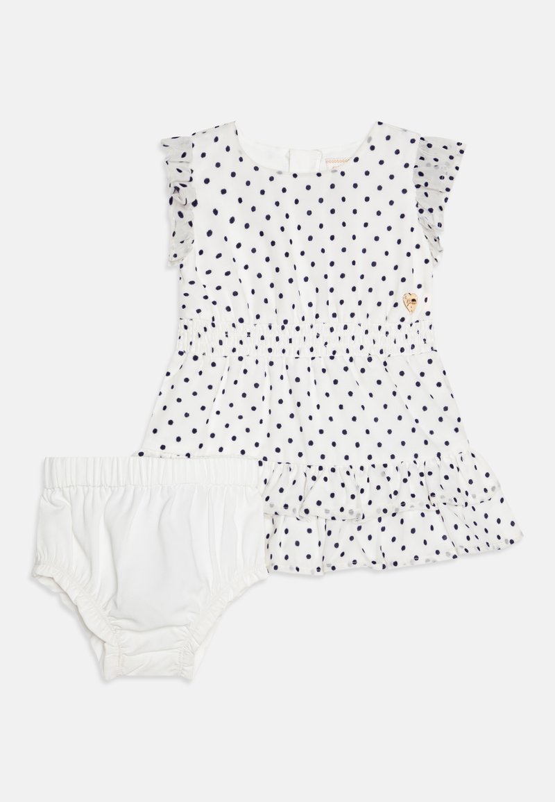 Balta medvilnės suknelė su tamsiai mėlynomis taškeliais, puoštomis volanėlėmis, ir sluoksniuota sijonėliu, derinama su atitinkamomis baltomis puošnimis.