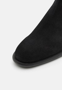 Bottine en daim noir avec un bout arrondi, une texture lisse et une fine semelle en caoutchouc. Aucun élément métallique ou accent visible.