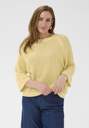 Kvinde med lysebrunt hår iført en løs, gul striktrøje og mørkeblå jeans, stående foran en ensfarvet baggrund.