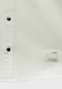 Jasno beżowa koszula z czarnymi guzikami i metką z informacjami o marce wszytą w lewym dolnym rogu. Tkanina ma gładką teksturę.