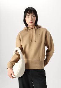 Beige mikina s kapucí, s elastickými lemy na rukávech a spodním okraji, s malým logem. Model drží bílou tašku s hladkým povrchem.