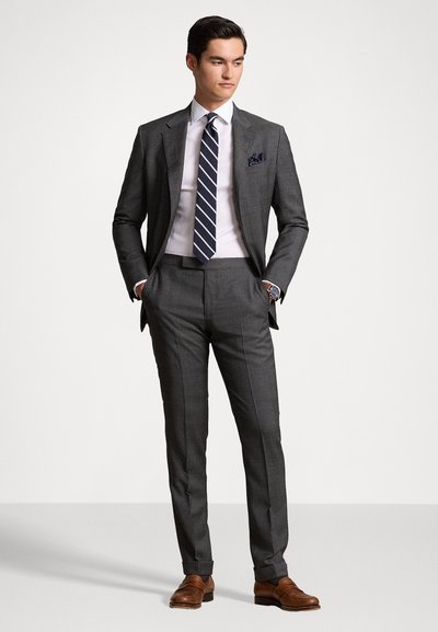 Costume gris sur mesure à motif tartan, chemise blanche et cravate rayée marine. Des chaussures en cuir marron et un mouchoir de poche marine complètent le look.