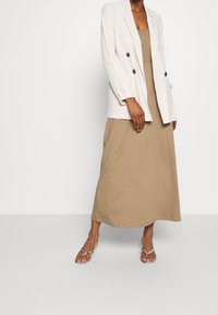 Žena oblečená v béžových maxi šatech, bílém dvouřadém blejzru s hnědými knoflíky a sandálech se vzorem hadí kůže, stojící na jednobarevném pozadí.