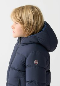 Giubbotto imbottito blu navy con cappuccio, con texture trapuntata, apertura con zip e una toppa circolare sul braccio sinistro.