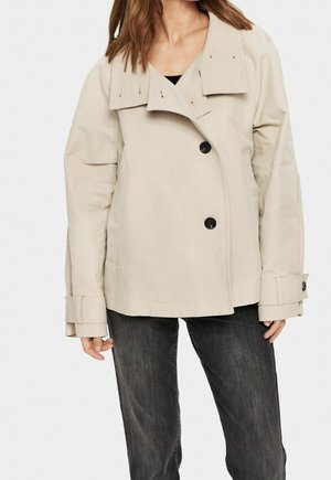 Kvinde iført en beige dobbeltradet jakke med stor krave og sorte knapper, kombineret med mørkegrå jeans.